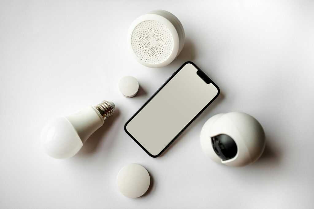 Flat lay of smartphone and smart home devices on a white backdrop, showcasing modern technology.